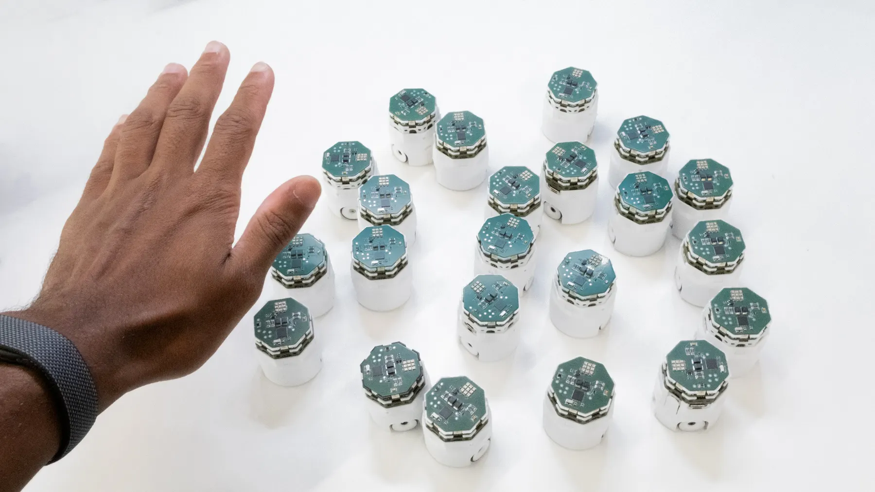 A hand hovering above a swarm of small white robots with green circuit-board tops.